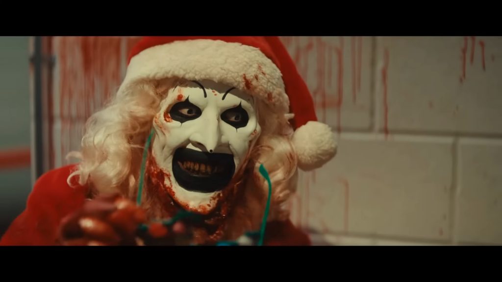 A menacing Art the Clown, wearing a Santa Claus hat and costume, smiling dementedly while standing inside a tiled shower in the Christmas horror sequel Terrifier 3