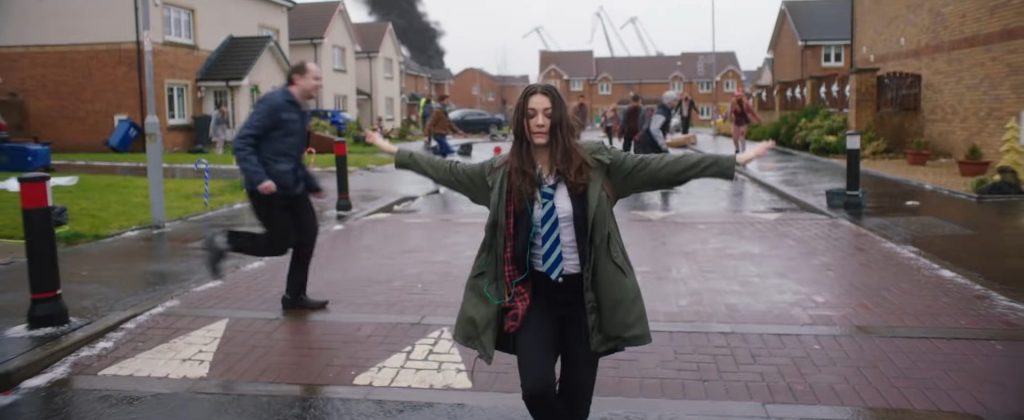 Ella Hunt as Anna dancing joyfully down a sunny suburban street, completely oblivious to the zombie apocalypse chaos and violent attacks happening in the background behind her in the festive musical horror comedy Anna and the Apocalypse.