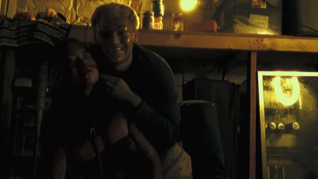 A chilling scene from the found footage horror film "Milk & Serial" (2024), showing a man in a translucent mask grinning while holding a victim in a dimly lit, cluttered basement or workshop.