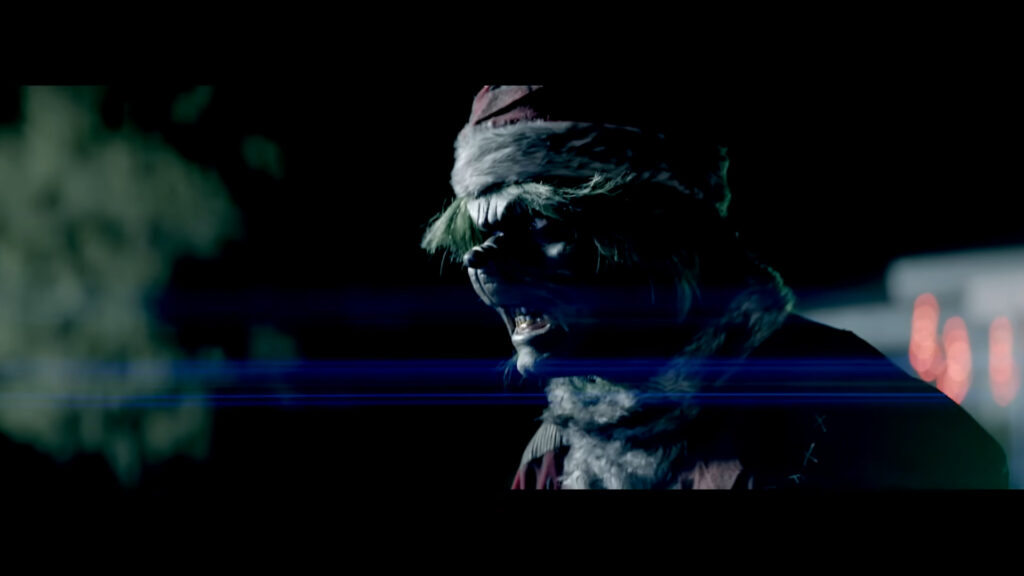 A menacing, monstrous version of a Christmas creature wearing a Santa hat and snarls in the dark in the horror movie The Mean One (2022).