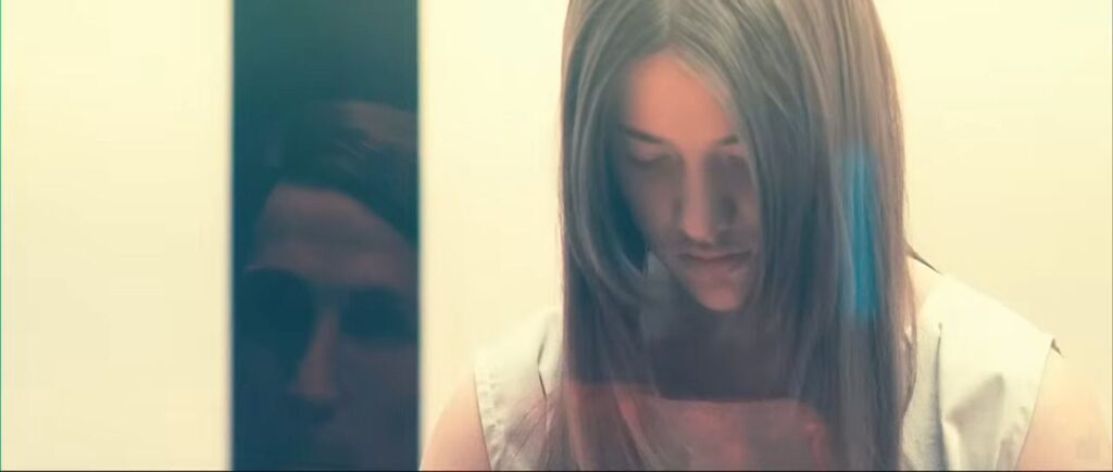 Beyond the Black Rainbow (2010): A woman in a sterile, white, retro-futuristic hallway with glowing light effects, capturing a cold liminal space aesthetic.