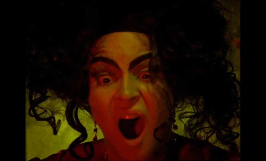 A close-up, high-contrast shot of a screaming character from the 2025 horror revival 'Dead Lover'. The character has exaggerated, dark arched eyebrows, curly black hair, and an open-mouthed expression of terror or rage, illuminated by eerie yellow and red cinematic lighting.