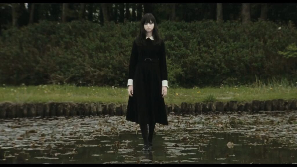 A young woman in a black dress with a white collar stands eerily still in the middle of a shallow, leaf-covered pond in the Japanese horror movie Fatal Frame.