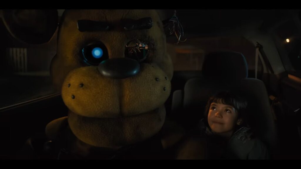 A large, weathered Golden Freddy animatronic with a glowing blue eye sits in a car next to a young girl in a scene from the Five Nights at Freddy's movie (2023).