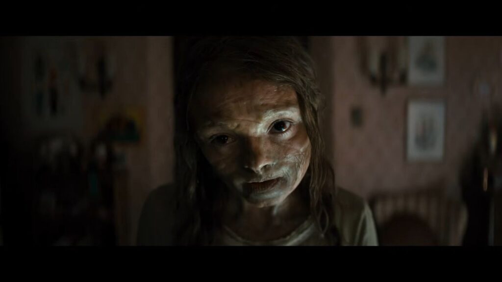 A haunting close-up of a character with pale, waxy, and textured skin from Lee Cronin’s 2026 'The Mummy'. The figure stares directly into the camera with dark, sunken eyes in a dimly lit, domestic setting, creating a modern and gritty reimagining of the classic horror theme.