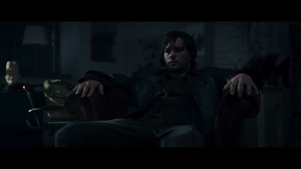 A man sits in a leather armchair in a dark, moody room, appearing exhausted in a still from the psychological horror film Return to Silent Hill.