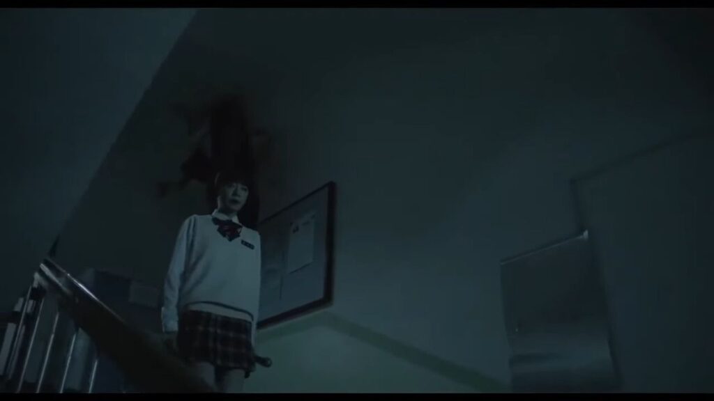A student in a school uniform stands on a dark staircase while a ghost-like figure hangs from the ceiling above her in the South Korean horror film The Labyrinth.