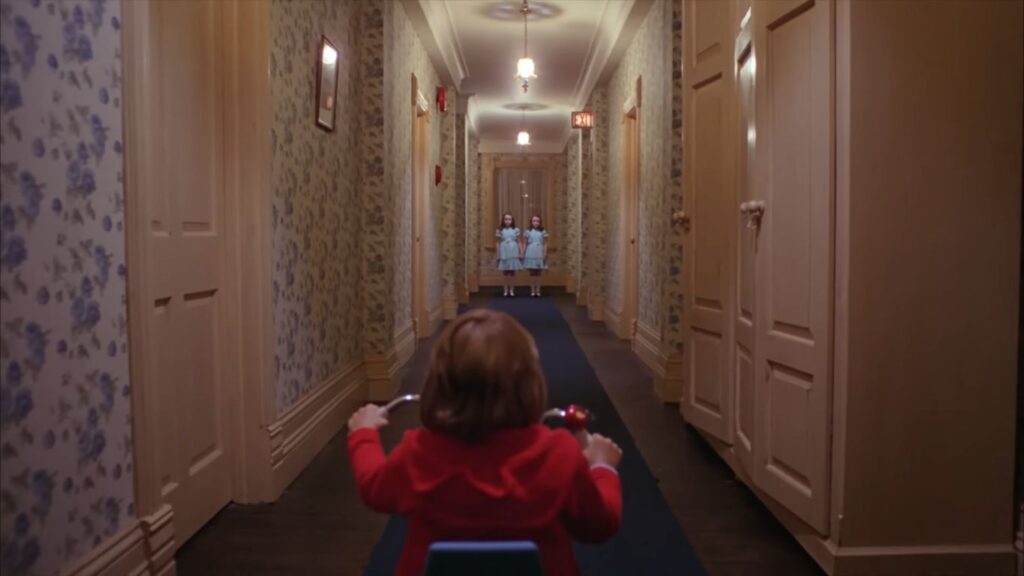 A young boy in a red sweater, seen from behind, rides a tricycle down a long hotel hallway with floral wallpaper. At the far end of the corridor, two identical twin girls in blue dresses stand side-by-side, staring directly at him in an eerie, symmetrical composition from The Shining.