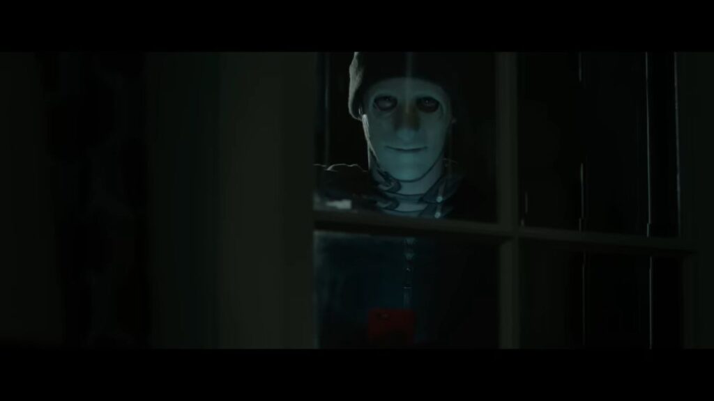 A masked killer wearing a beanie looks through a dark glass window pane while holding a phone in the home invasion horror movie Hush (2016).