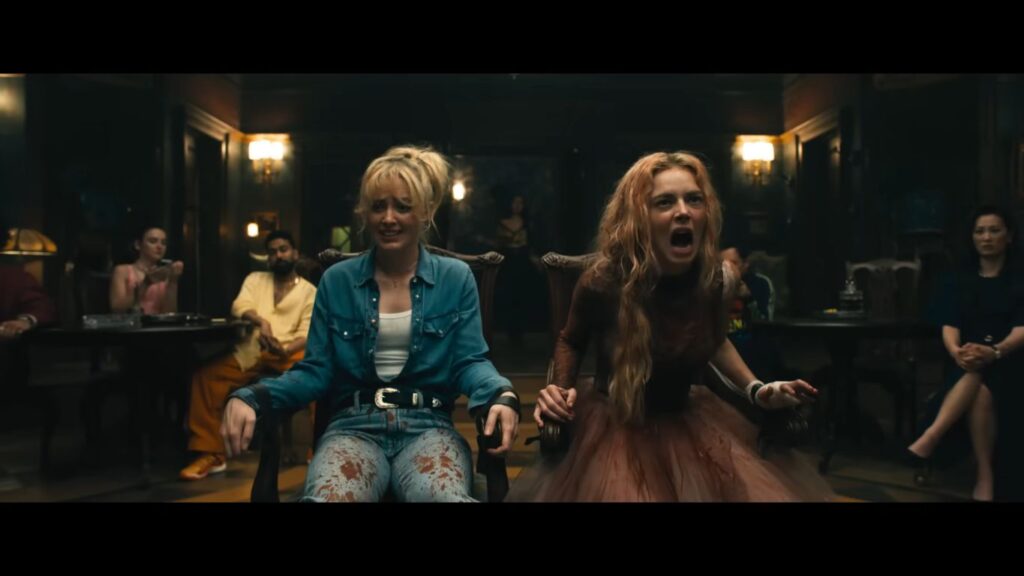 Two blood-splattered women sit tied to wooden chairs inside a mansion, with one screaming in terror or anger, from a badass horror movie scene.