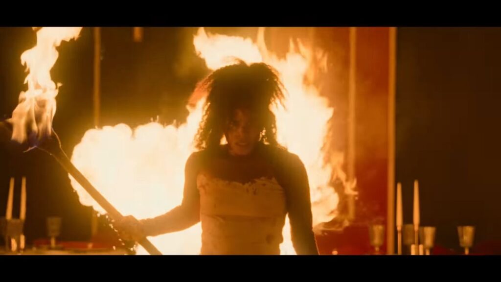 A fierce woman stands holding a blazing fire torch with massive flames exploding behind her in a dramatic scene from the horror movie They Will Kill You (2026).