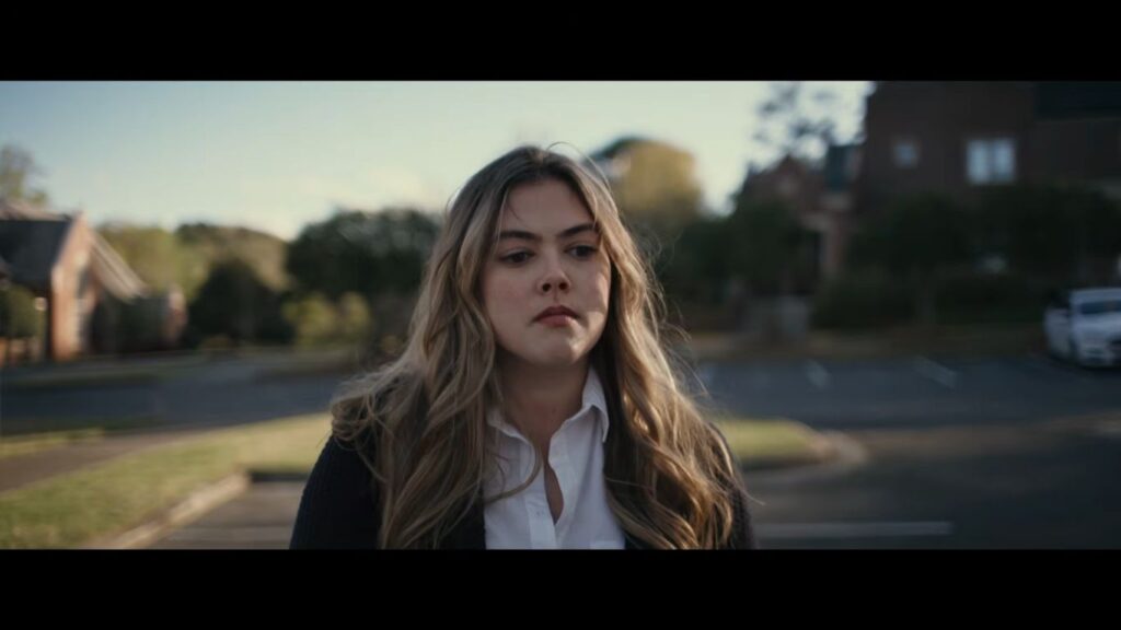 A determined young woman with long blonde hair wearing a white collared shirt walks through an outdoor parking lot in the horror thriller You're Killing Me (2023).