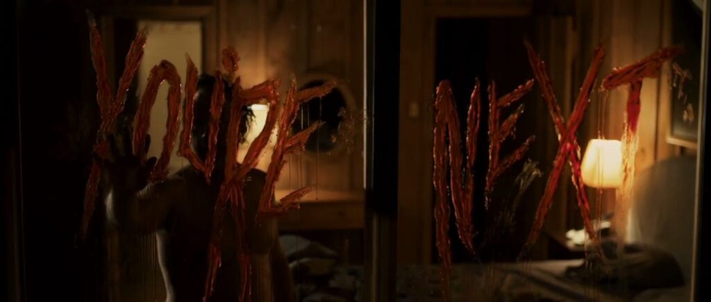 The words You're Next written in dripping blood across a glass window pane with a dark figure silhouetted behind it in the slasher horror movie You're Next (2011).