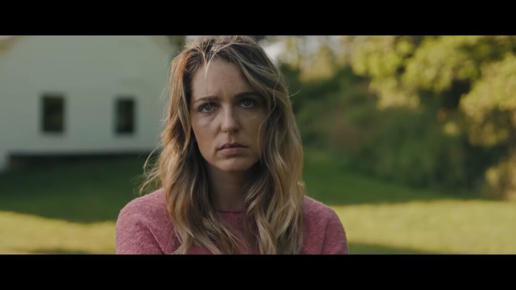 A serious-looking blonde woman in a pink sweater stares directly ahead in a close-up shot from the horror movie Affection (2025)