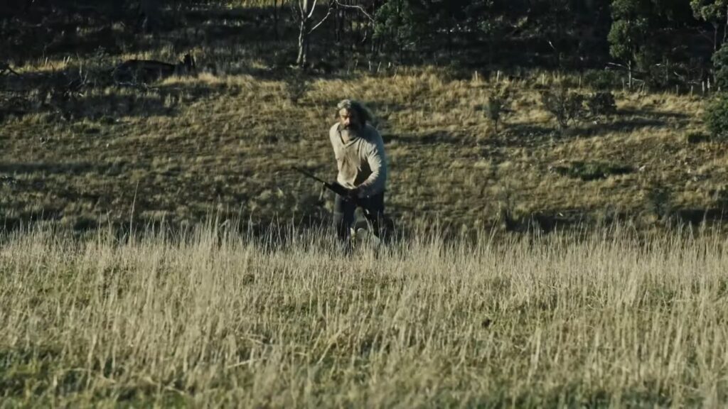 A bearded man holding a rifle running through a dry, grassy field in the Australian outback in Beaten to Death.