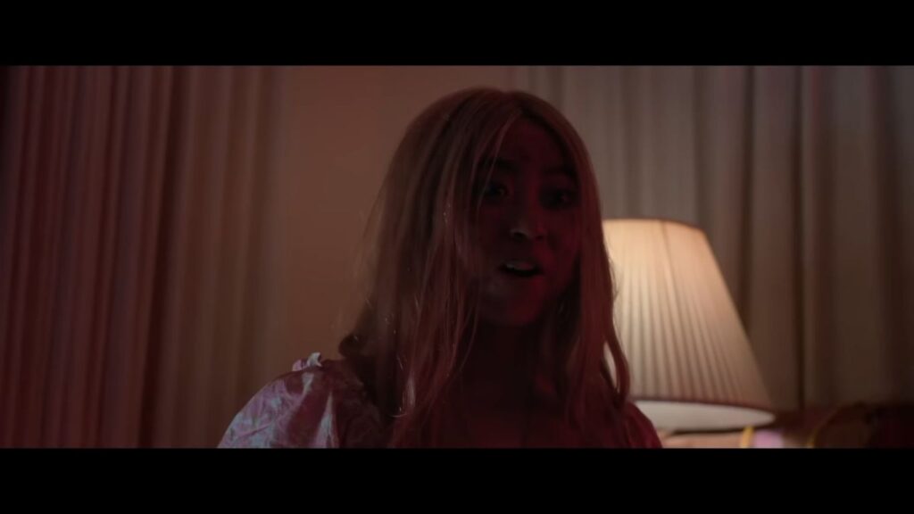 A young woman with long blonde hair and a pink top standing in a dimly lit, red-toned room in Grafted.