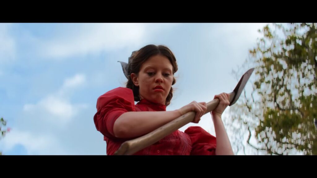 A low-angle shot of Mia Goth looking down intensely while gripping a large axe against a blue sky in the horror movie Pearl (2022).