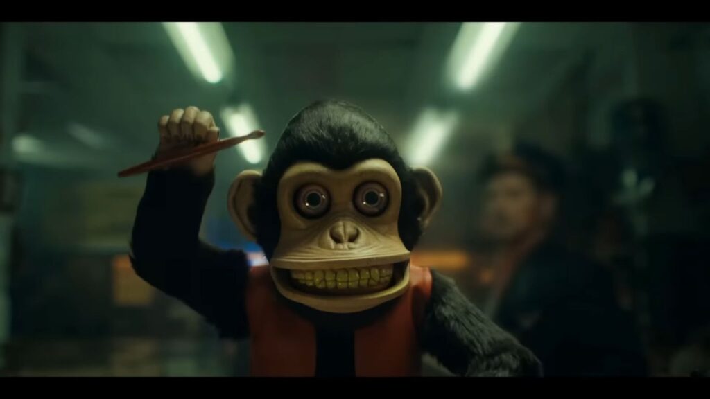 A menacing, wide-eyed mechanical toy monkey raising a drumstick with a sinister grin in The Monkey.