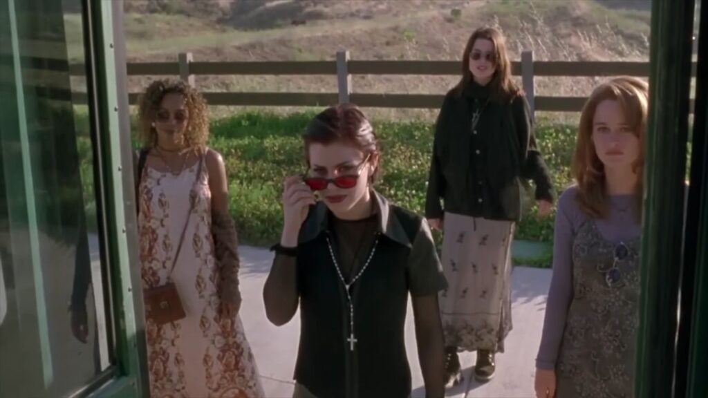 Fairuza Balk lowers her sunglasses to look into a camera while standing with her teenage coven outside a school bus in the supernatural horror movie The Craft (1996).