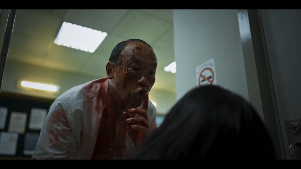 A blood-covered man with an unsettling expression leaning down and placing a finger to his lips in a shushing gesture in The Sadness.
